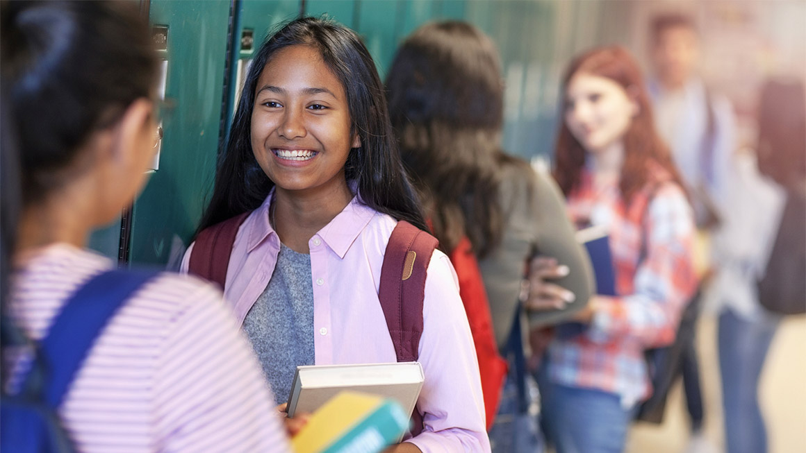 Cheerful teenage girls talking while standing at illuminated corridor. Female friends are by lockers in education building. Students are at high school.