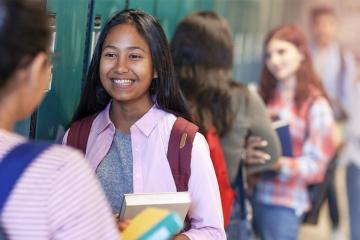 Cheerful teenage girls talking while standing at illuminated corridor. Female friends are by lockers in education building. Students are at high school.