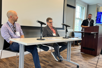 An image from a workshop eaturing two speakers at a table, and one person standing up. 